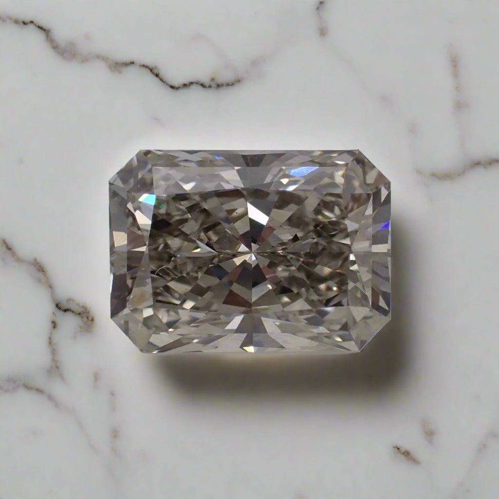 A loose, rectangular radiant cut diamond rests on a white surface. The stone has many, tiny inclusions giving it an overall grey appearance.