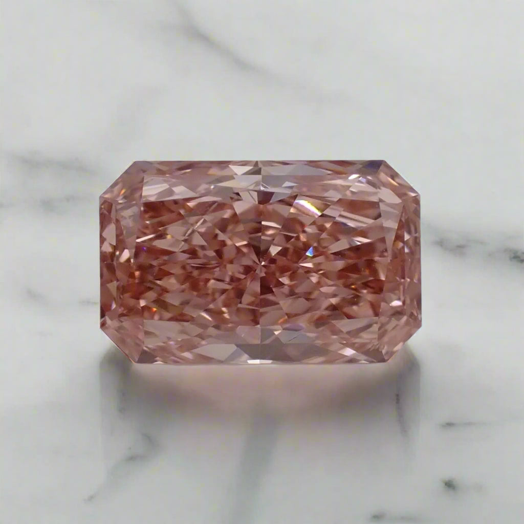 A radiant cut diamond rests on a white surface. The diamond is a bright, soft pink colour.