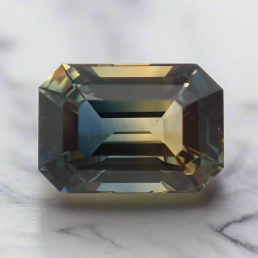 A rectangular sapphire rests on a white surface. The stone shows distinct patches of blue and peachy yellow.