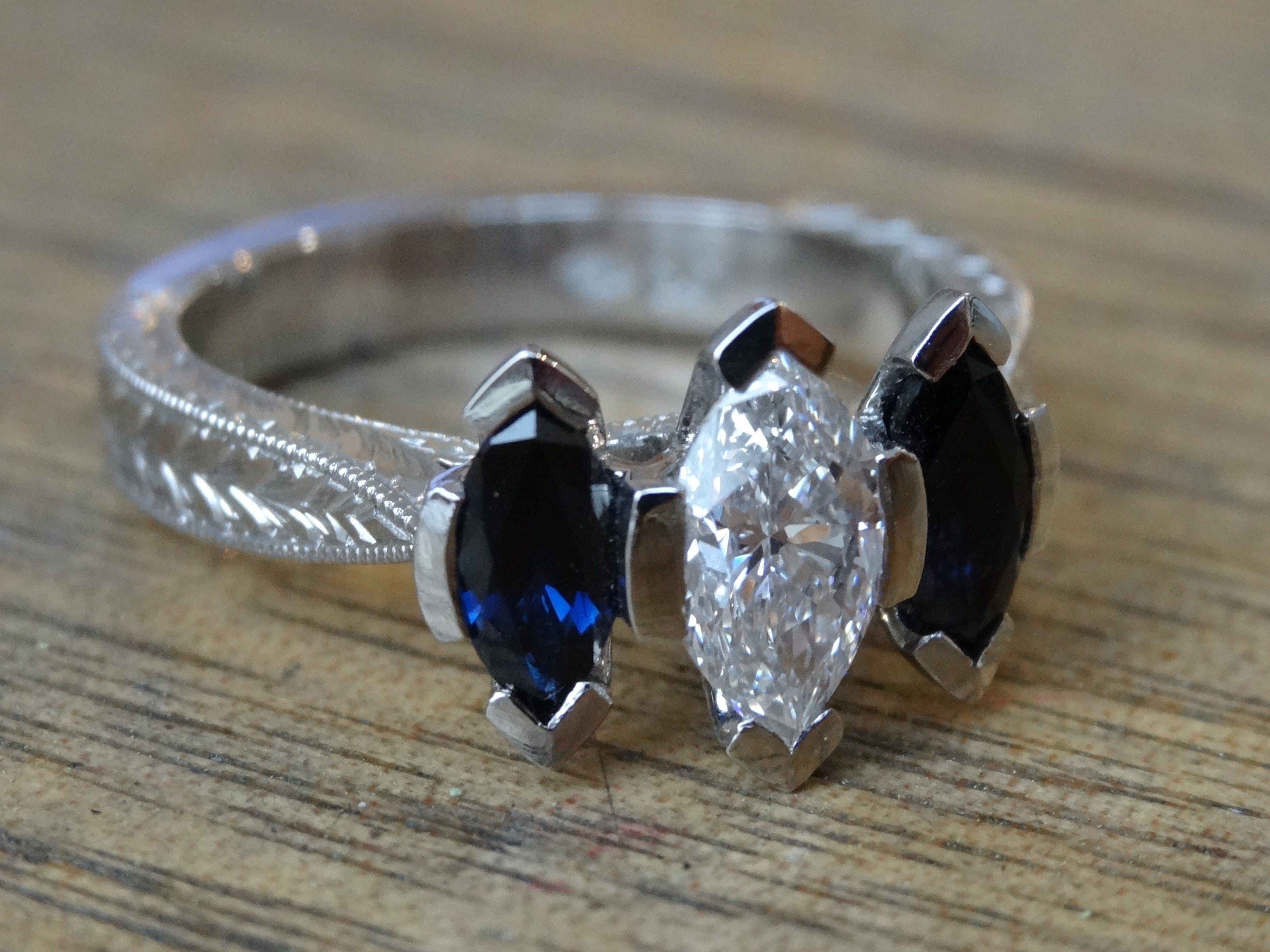 A trilogy engagement ring with a marquise cut diamond and pair of marquise cut sapphires is set in a hand engraved band.