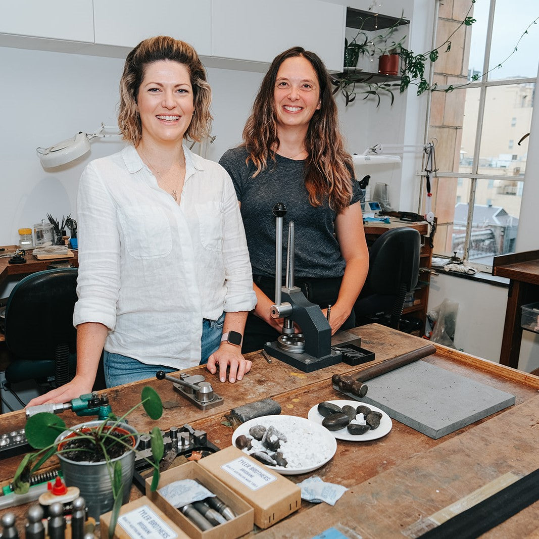 Jenna and Megan standing together behind a large wooden bench with assorted tools in their Melbourne jewellery studio.