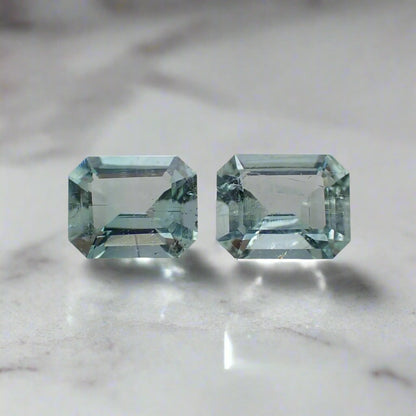 A pair of pale blue-green emeralds with visible inclusions rest on a white surface.