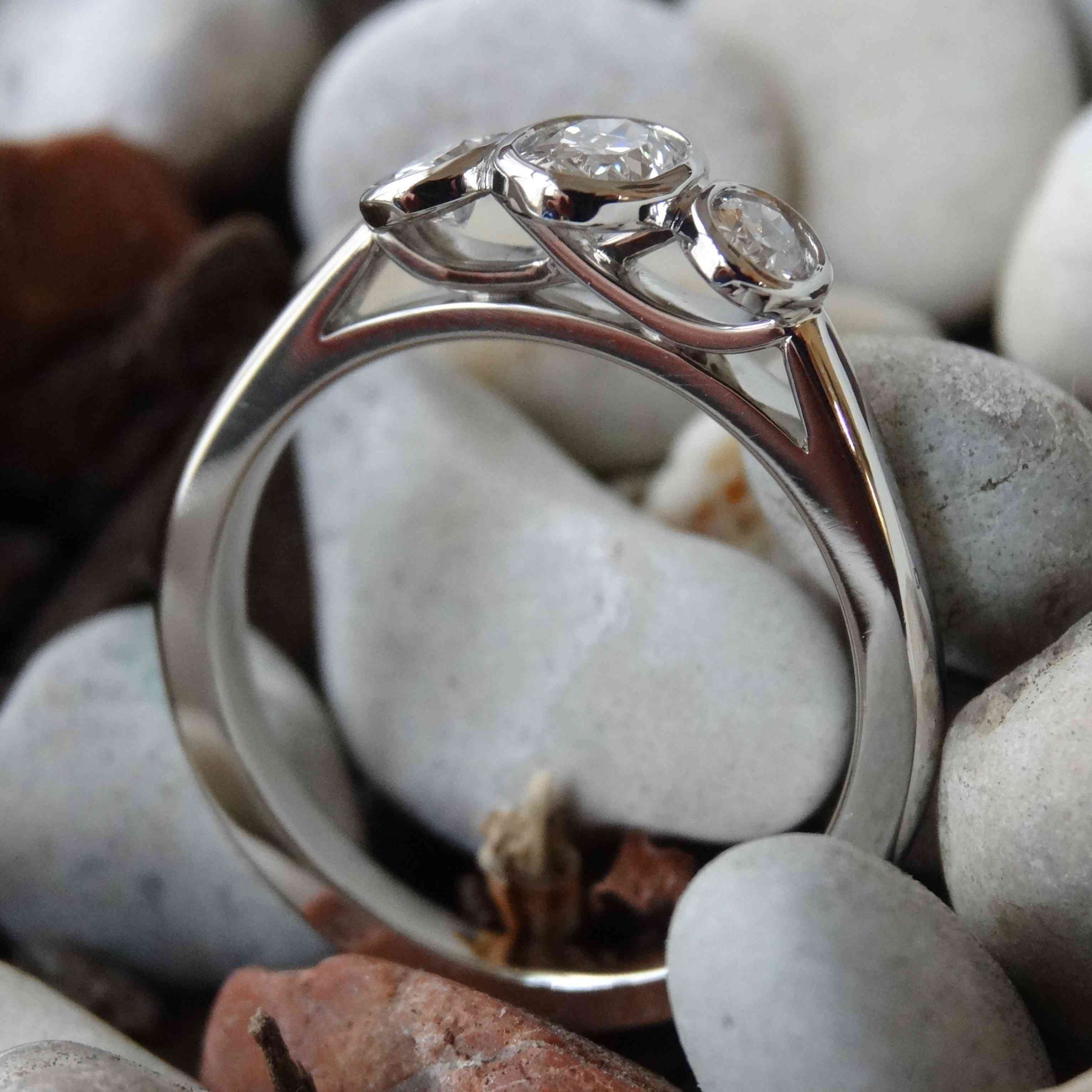 A platinum three stone engagement ring with unique side view with a backdrop of grey pebbles