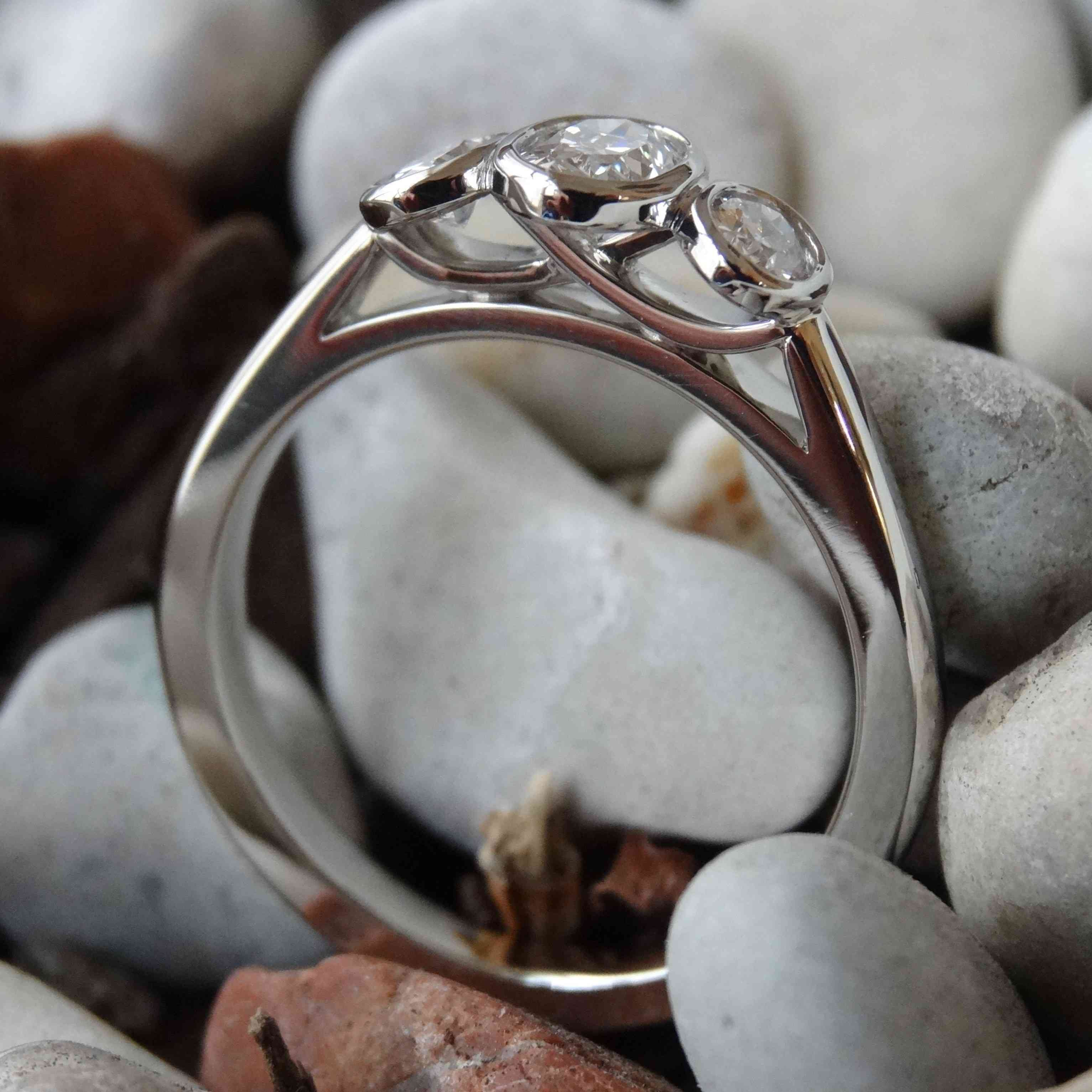 A platinum three stone engagement ring with unique cross over detail rests on a backdrop of grey pebbles.