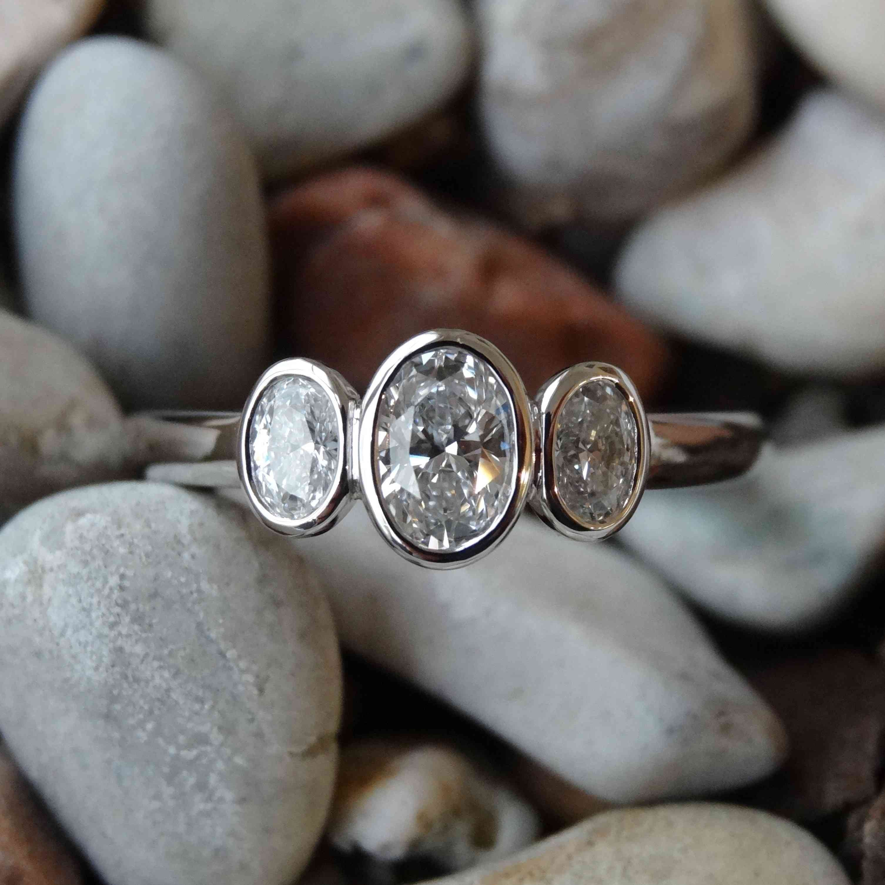 Platinum trilogy engagement ring bezel set with three oval diamonds laying on small grey pebbles.