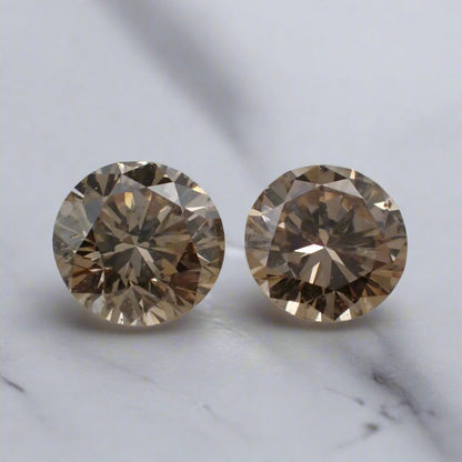 A pair of champagne, round brilliant cut diamonds rests on a white surface.