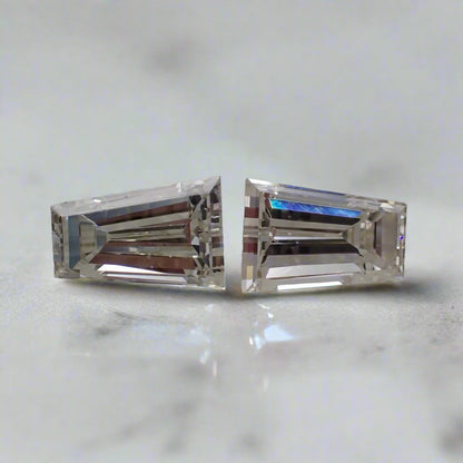 A pair of tapered baguette diamonds rest beside each other on a white surface.