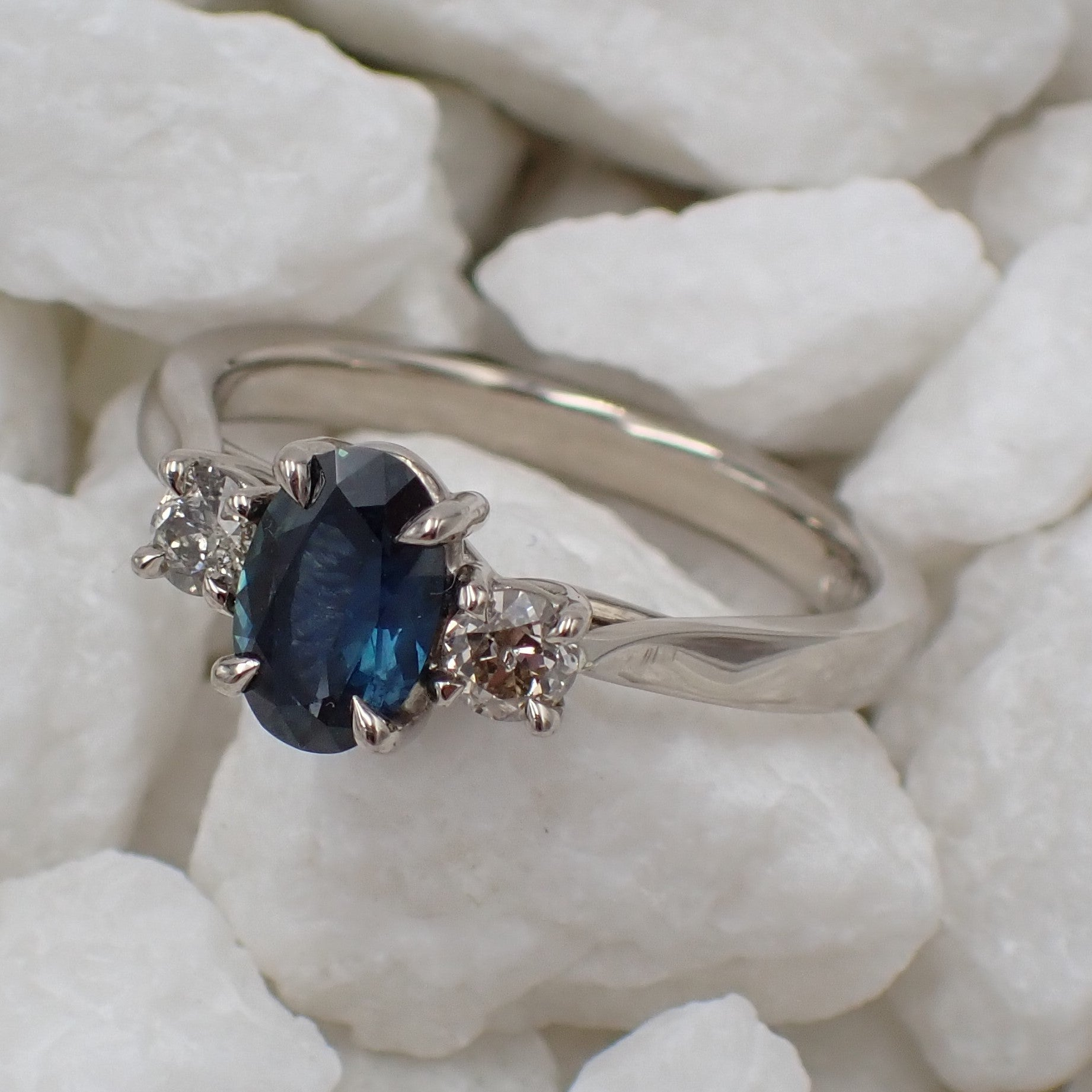 A blue oval sapphire and diamond claw set engagement ring in white gold lies flat on some rocks.