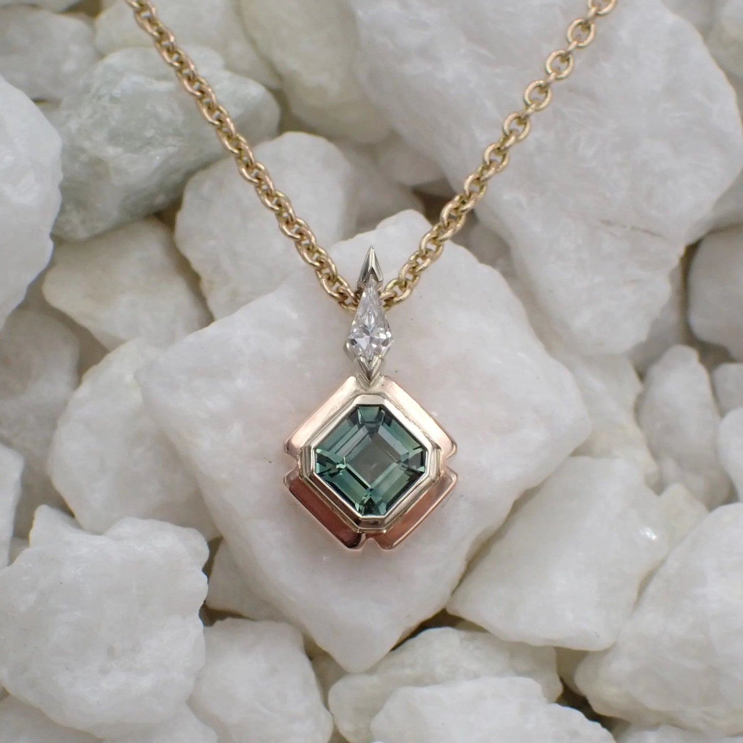 A light green asscher cut sapphire is bezel set in white gold with a rose gold outer bezel. A kite shaped diamond sits atop, set in white gold, with a chain running behind.