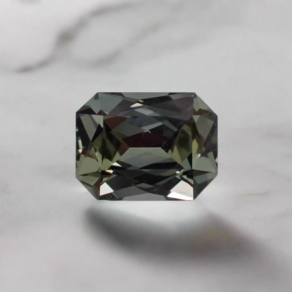 a grey, rectangular sapphire sits loose on a white surface. There are greenish flashes showing through the stone.