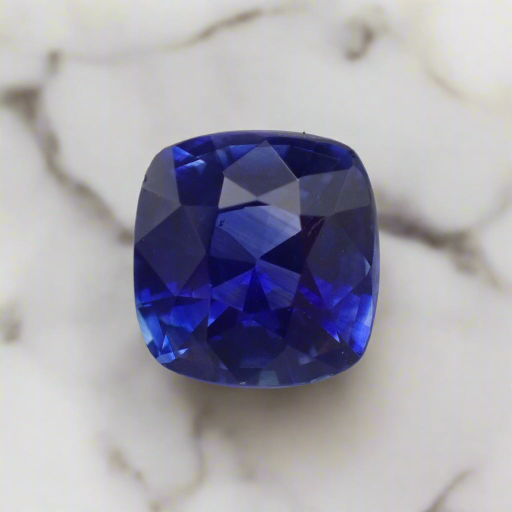 A very vibrant blue sapphire rests on white marble. The stone is a cushion cut, almost square.