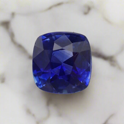 A very vibrant blue sapphire rests on white marble. The stone is a cushion cut, almost square.