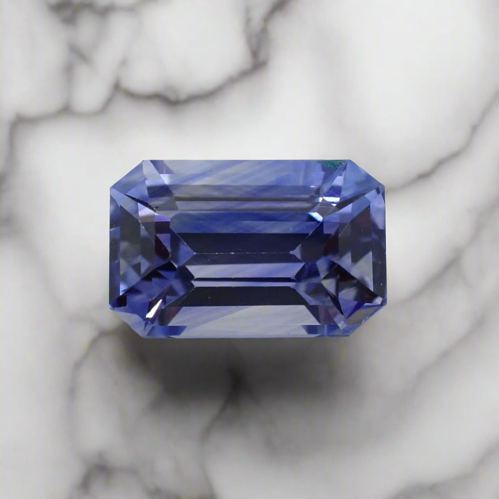 An emerald cut sapphire rests on white marble. The sapphire shows banding of light purplish blue and white.