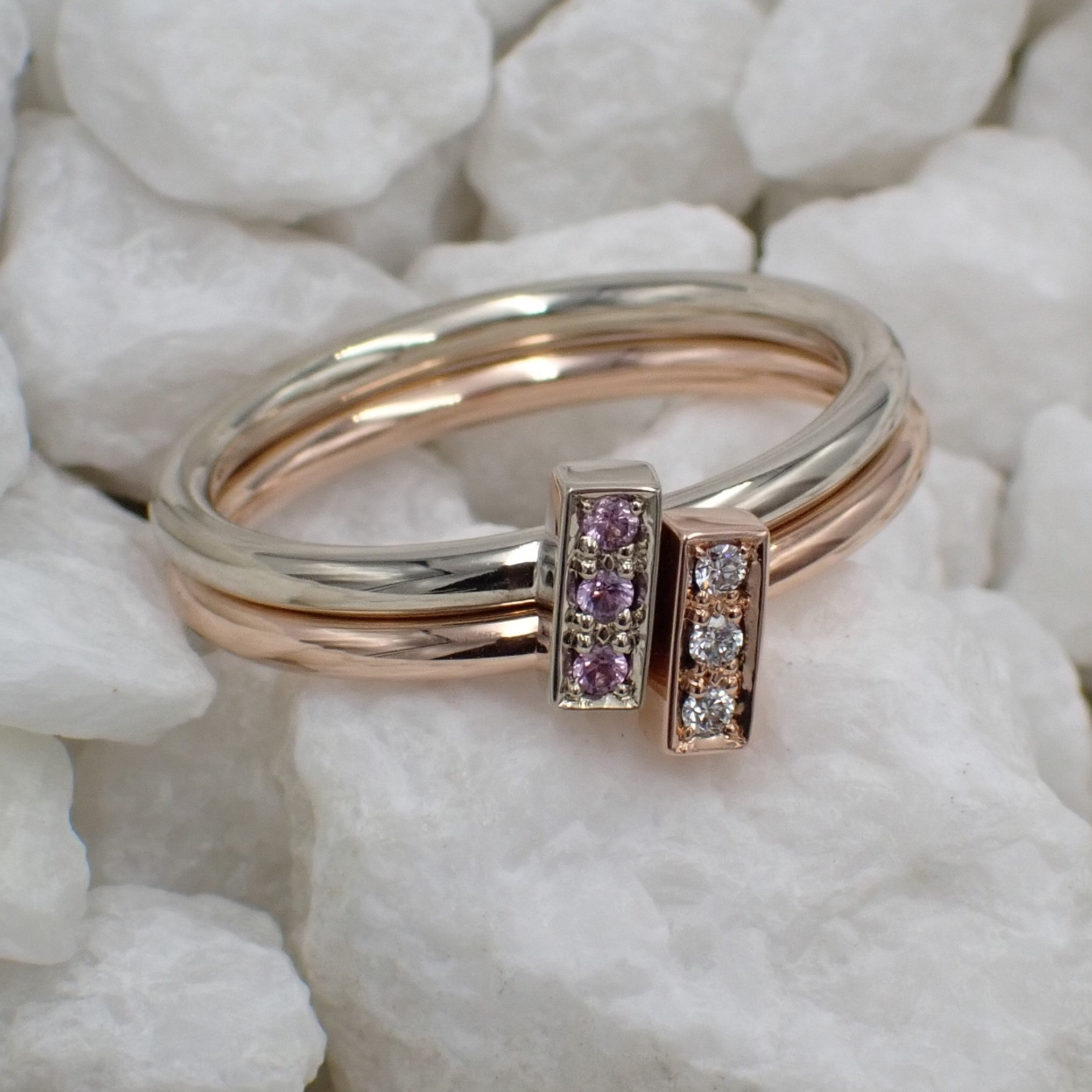 Two geometric stackable rings rest on white rocks. One is white gold with pink sapphires and the other is rose gold with white diamonds.