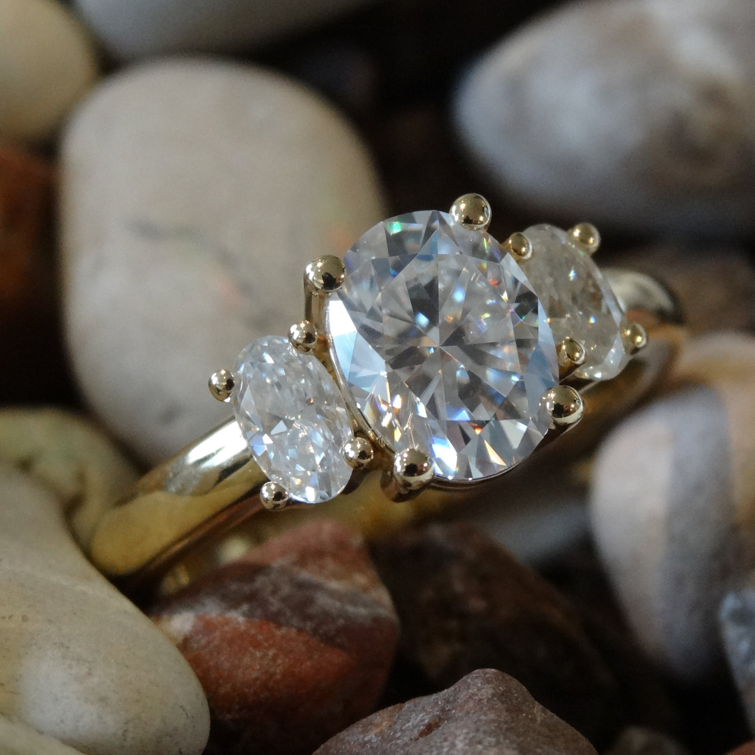 Three oval cut moissanites are claw set into a yellow gold engagement ring. The ring is resting on multi coloured pebbles.