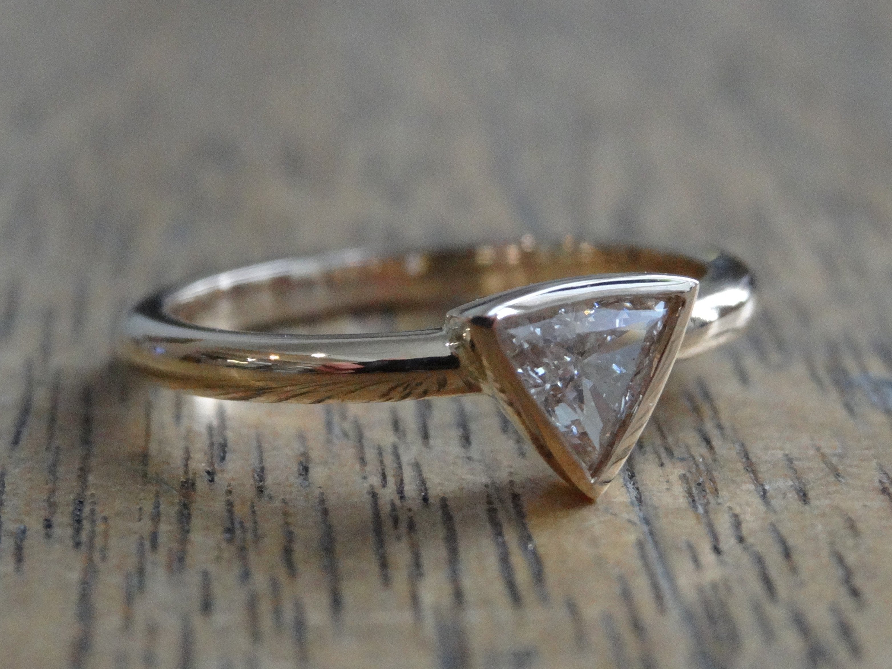 A trilliant cut diamond engagement ring, bezel set, with a narrow band, on a timber table top.