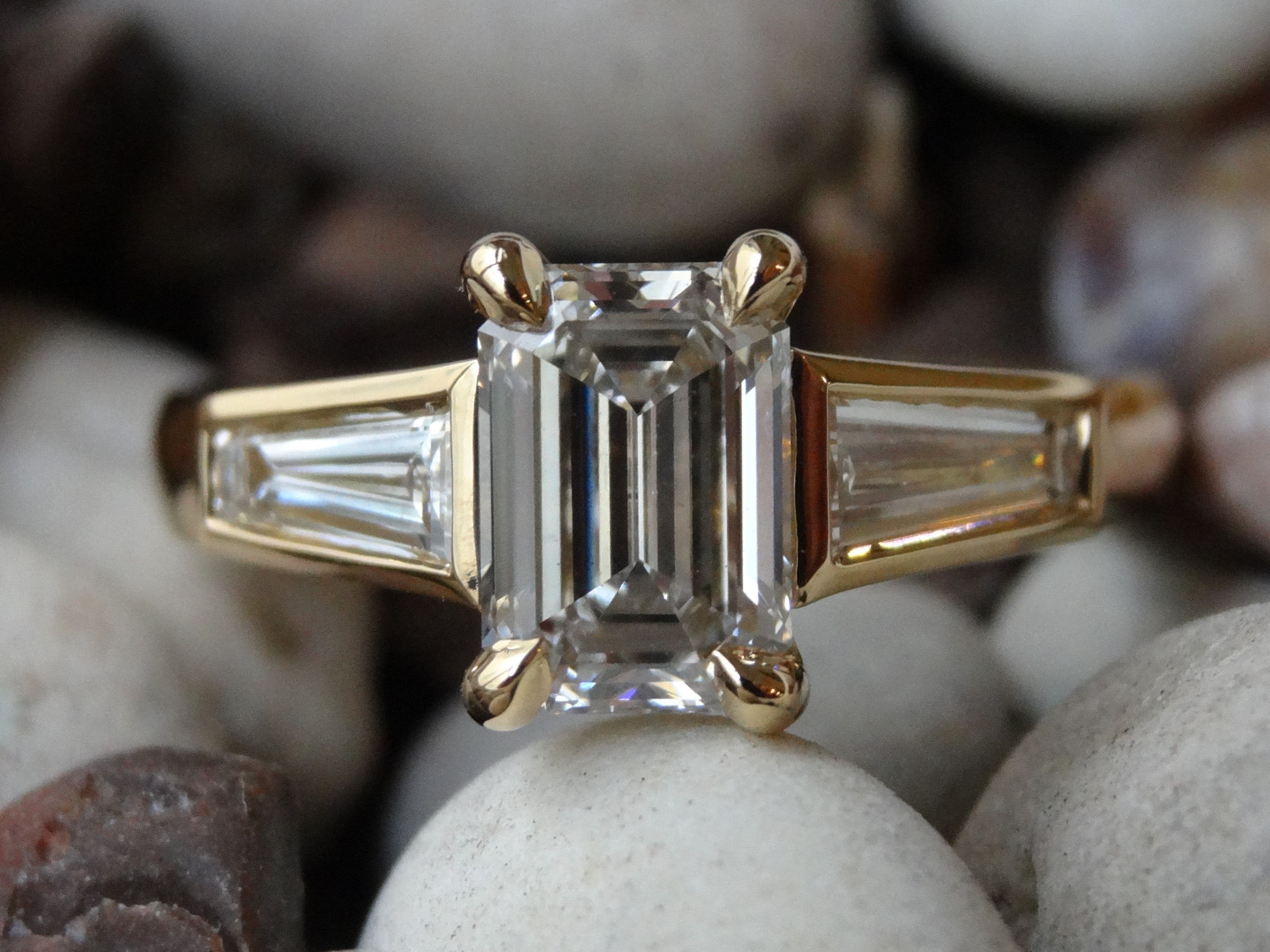 A yellow gold engagement ring, with a claw set emerald cut diamond and bezel set pair of tapered baguettes rests on pebbles.