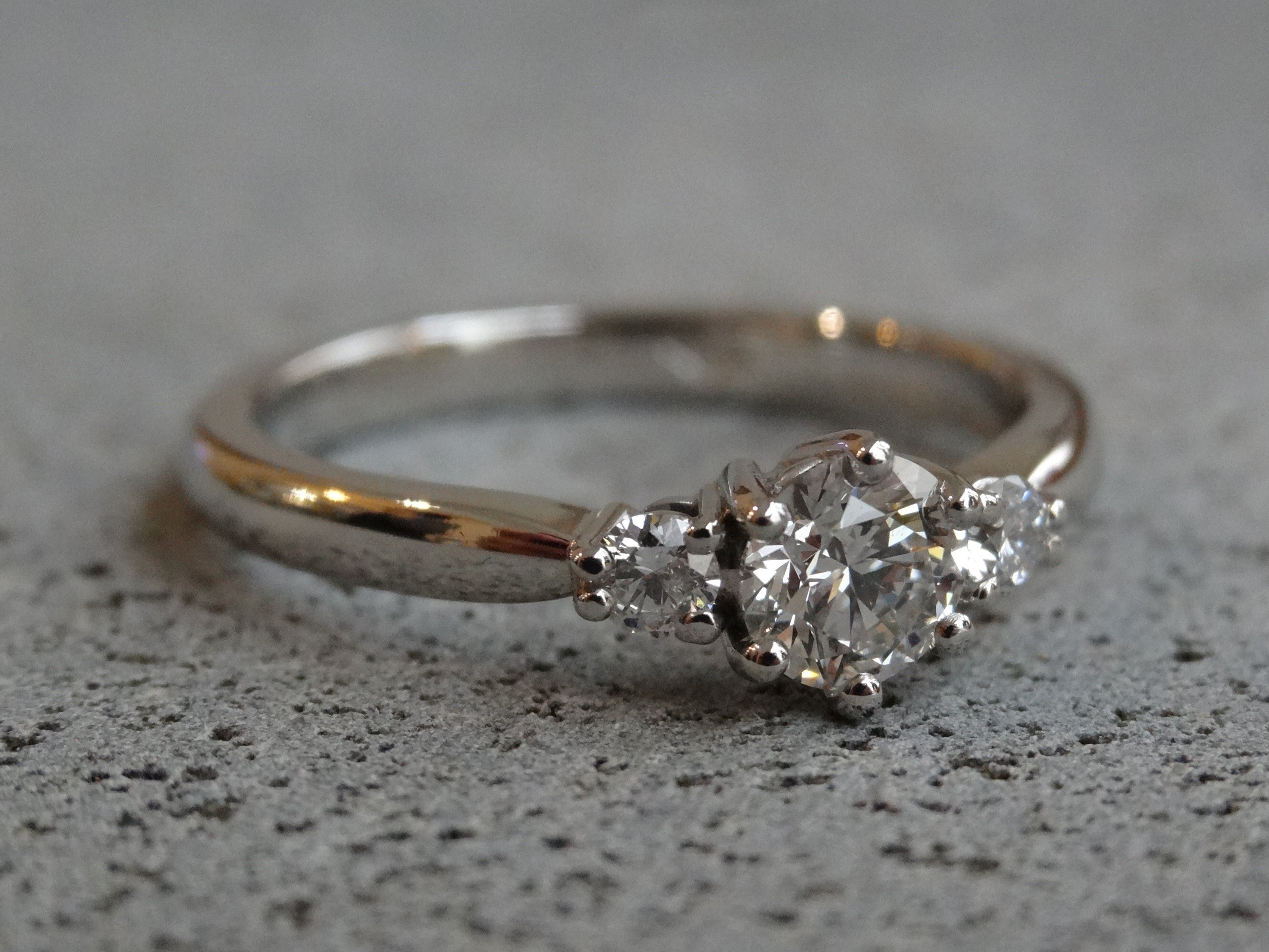 A trilogy engagement ring, in platinum, with round diamonds rests on a rough concrete surface.