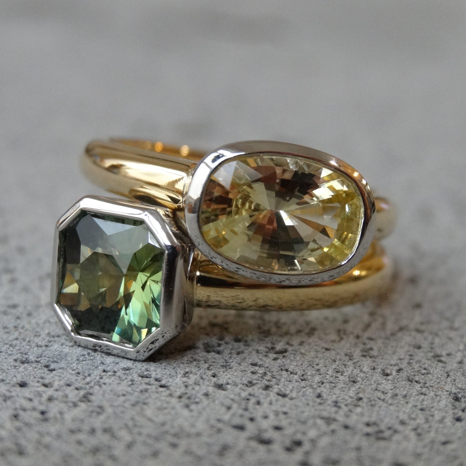 18ct yellow and white gold bezel set oval yellow sapphire solitaire ring sitting on top of a radiant shaped green sapphire bezel set solitaire.