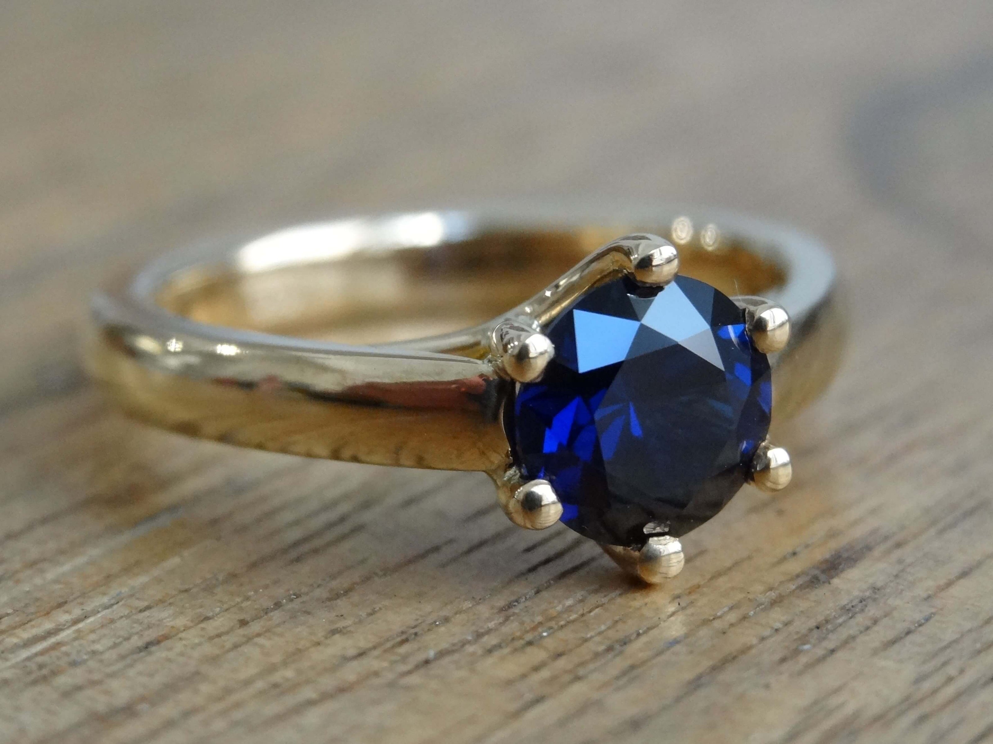 18ct yellow gold 6 claw engagement ring set with one round bright blue sapphire on a wooden surface 